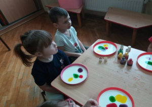 dzieci siedzą przy stolikach ia na talerzach z mlekiem maja kolorowe plamy